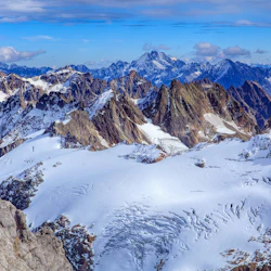 Snötäckta bergstoppar under en klarblå himmel med spridda moln. Tuff terräng med steniga utgrävningar.