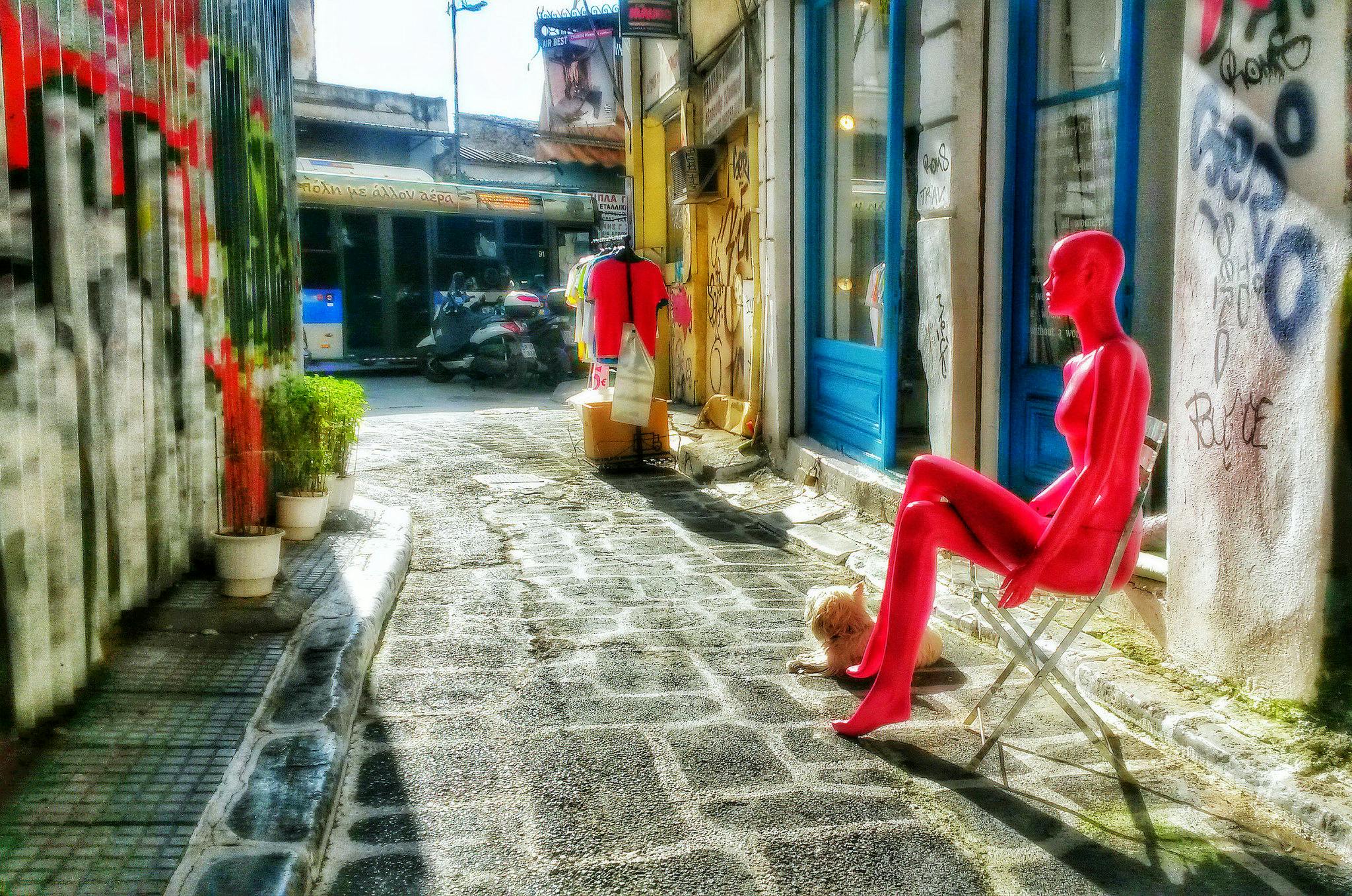 En smal gata med graffititäckta väggar, färgglada skyltdockor, en liten hund och en butik med blå fönster.