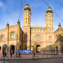En stor, utsmyckad byggnad med randiga väggar och dubbla kupolformade torn, människor som går i närheten, under en klarblå himmel.