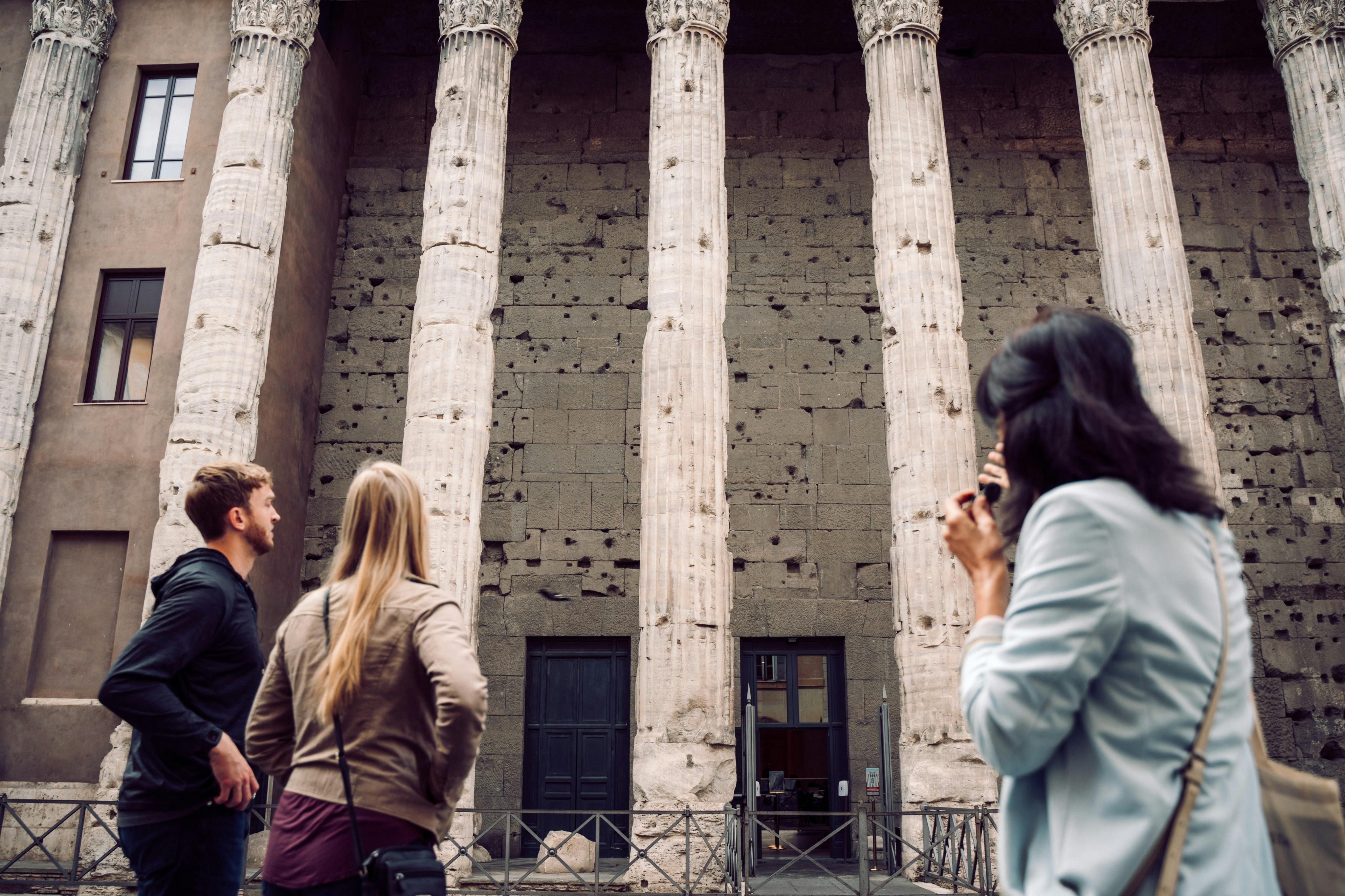 Due persone si trovano di fronte a un antico edificio con colonne alte e consumate e un muro di pietra strutturato.