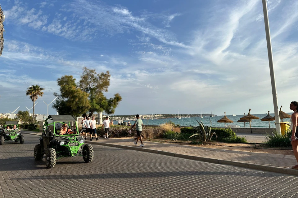 People walking near a beachfront with a pathway, a green dune buggy, trees, umbrellas, and the sea under a partly cloudy sky.