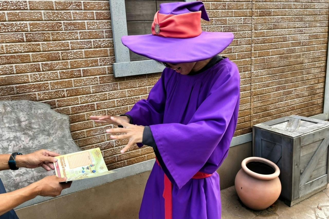 A person in a purple robe and hat, resembling a wizard, gesturing dramatically near a brick wall and a pottery item.