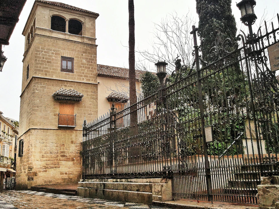 Stone building with barred windows next to an ornate black iron fence, cobblestone pathway, and tall trees in the background.