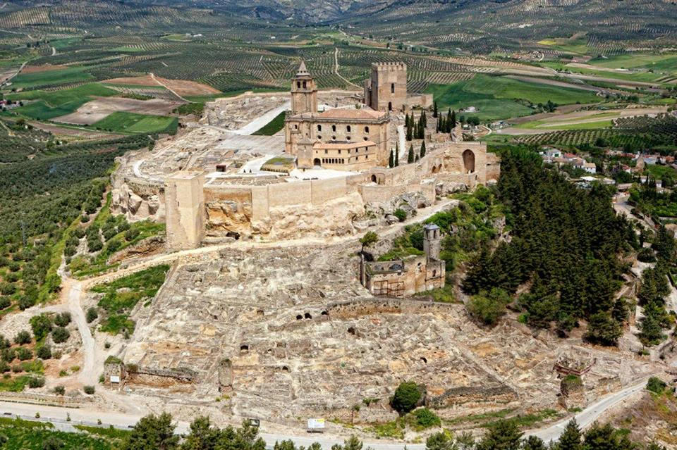 An aerial view of a historic stone castle and the surrounding ruins, set amidst a lush, green, hilly landscape.