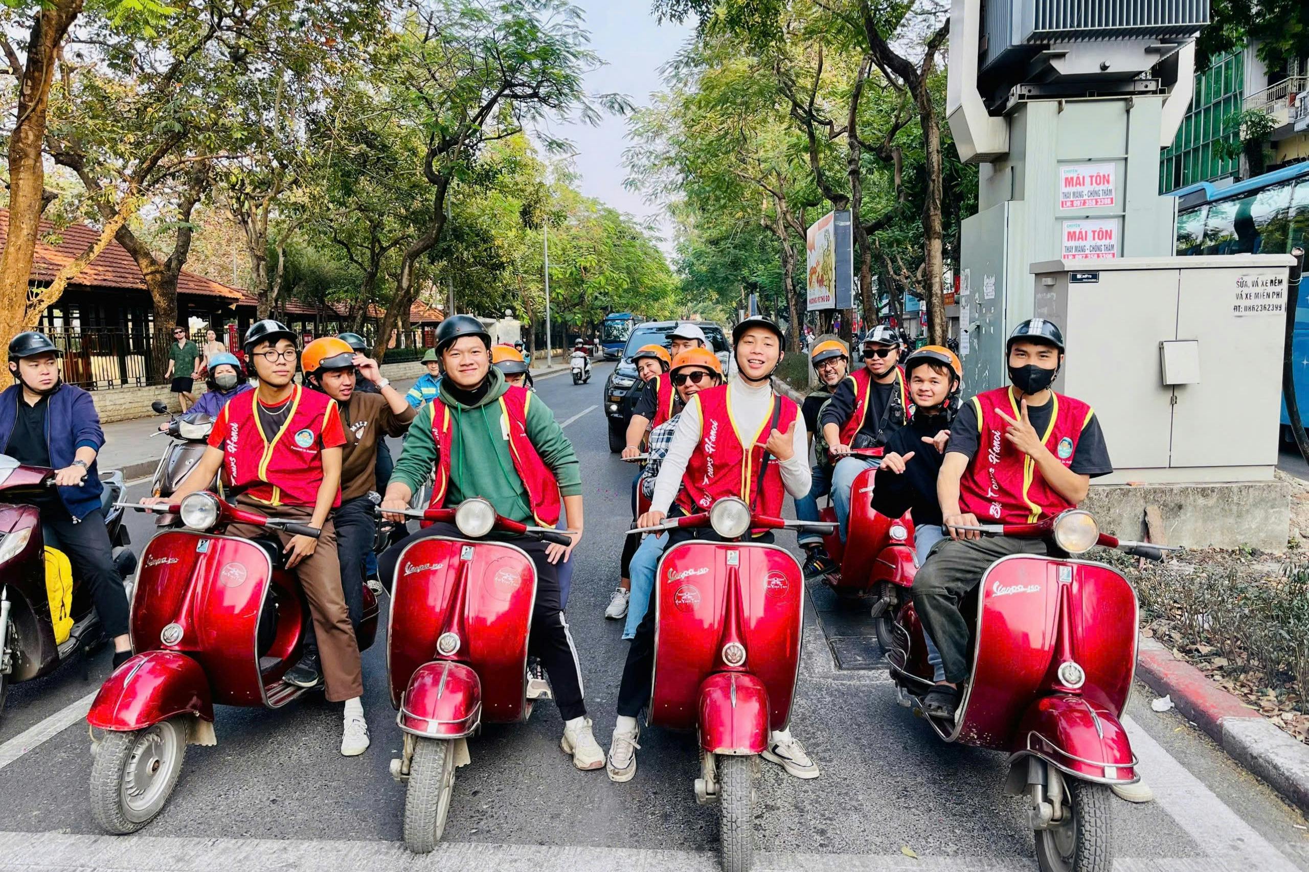 Group of people in helmets and red vests on red scooters, smiling and posing on a tree-lined street.