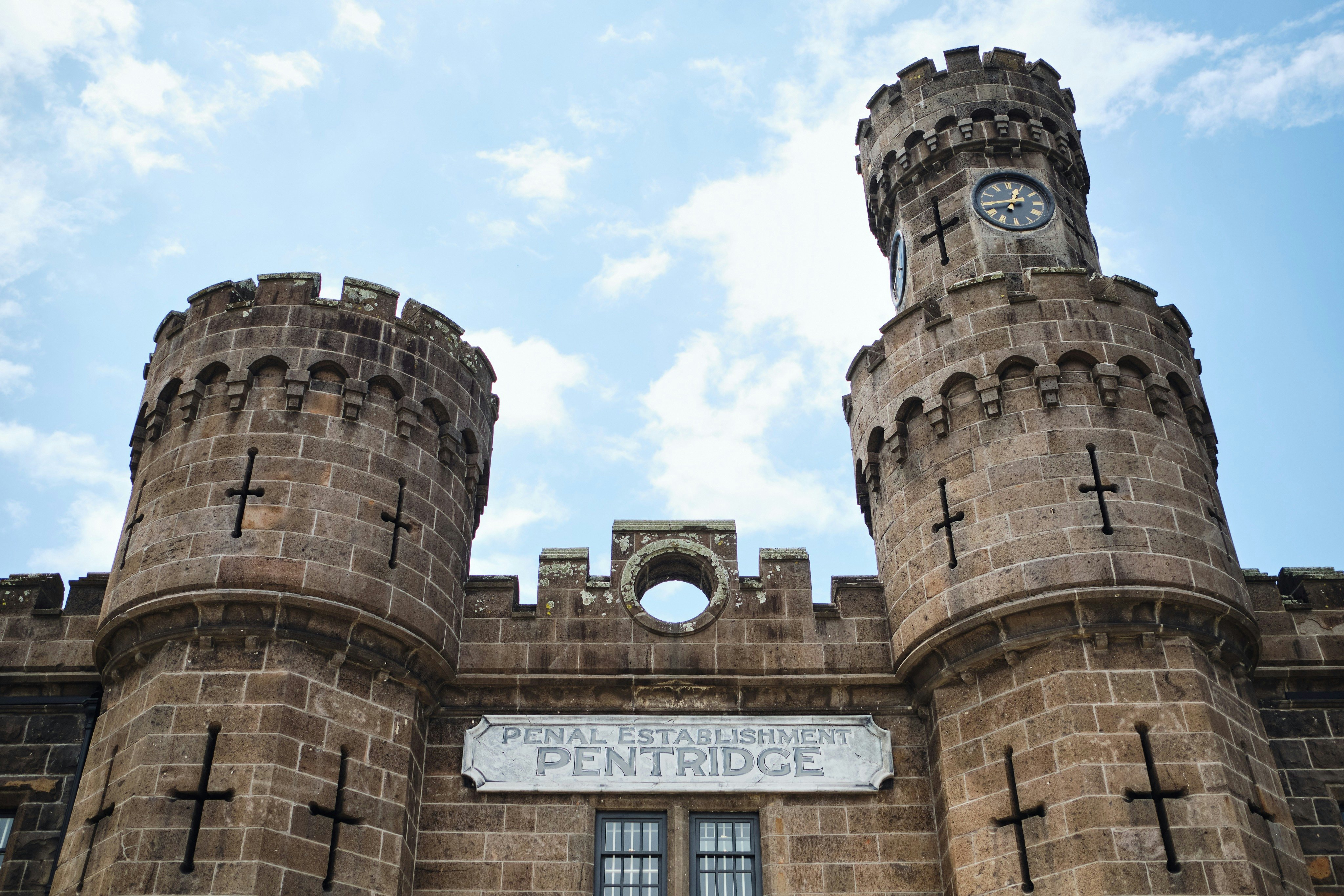 Edifício de pedra com duas torres redondas, uma das quais com um relógio. O letreiro diz "Estabelecimento Penal de Pentridge". Céu azul com nuvens.