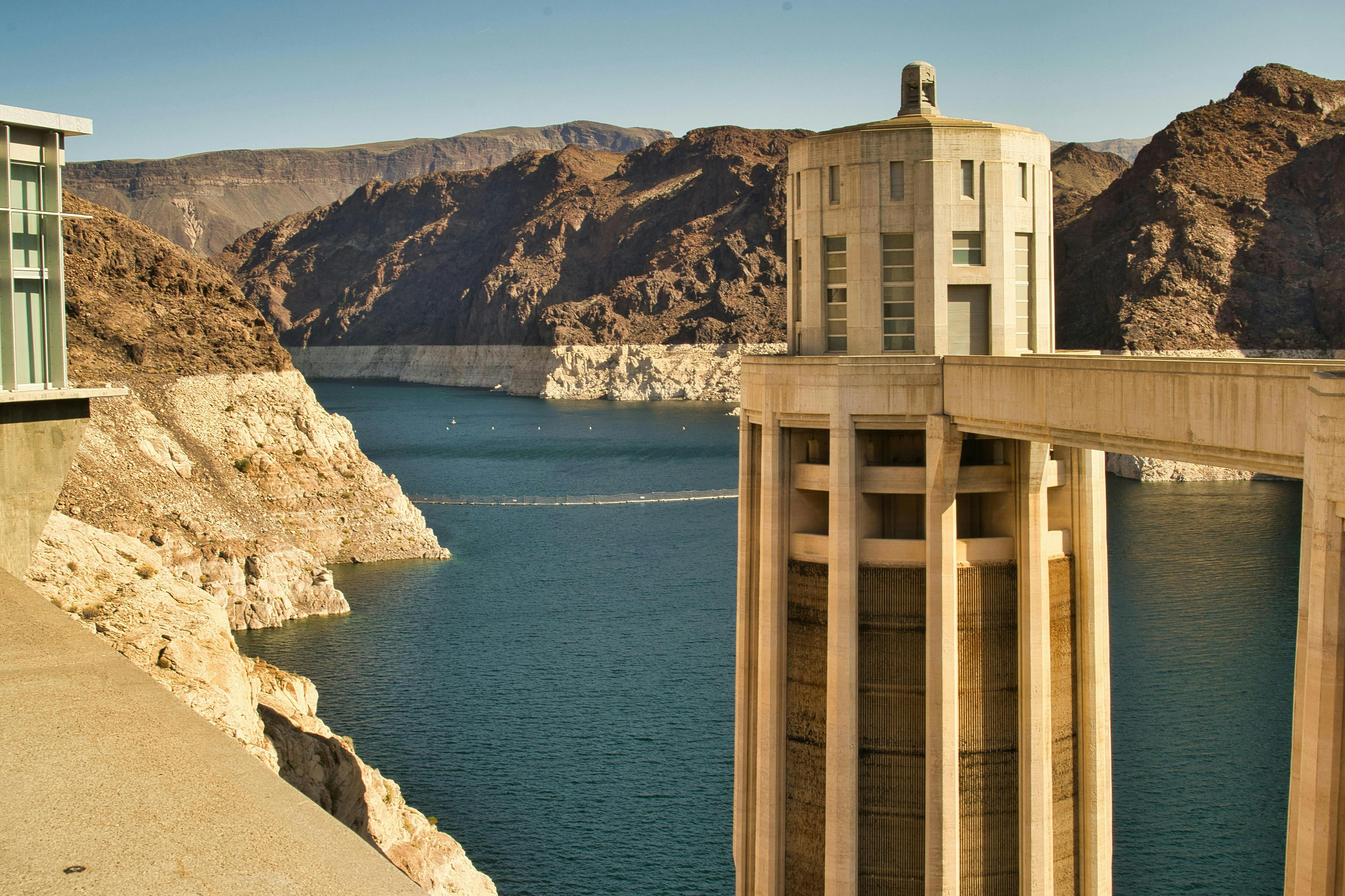 A view of a dam tower structure with a lake and rugged mountains in the background under a clear sky.