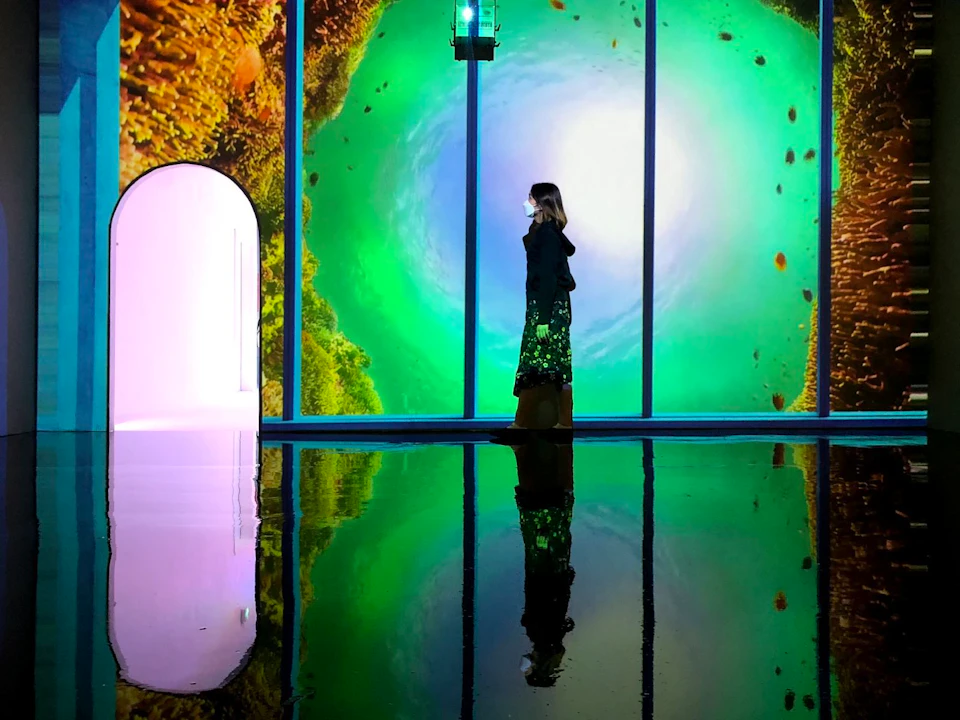A person stands in a reflective room with vibrant green and yellow wall projections and a glowing arched doorway.