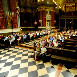 En stor, utsmyckad kyrklig interiör med högt i tak och många människor sittande i bänkar, vänd mot framsidan.
