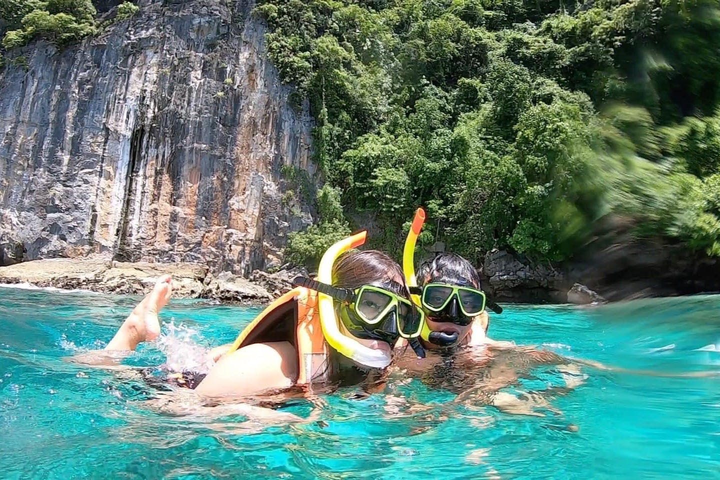 两个人在清澈的绿松石海水中浮潜，靠近多岩石的森林海岸线。