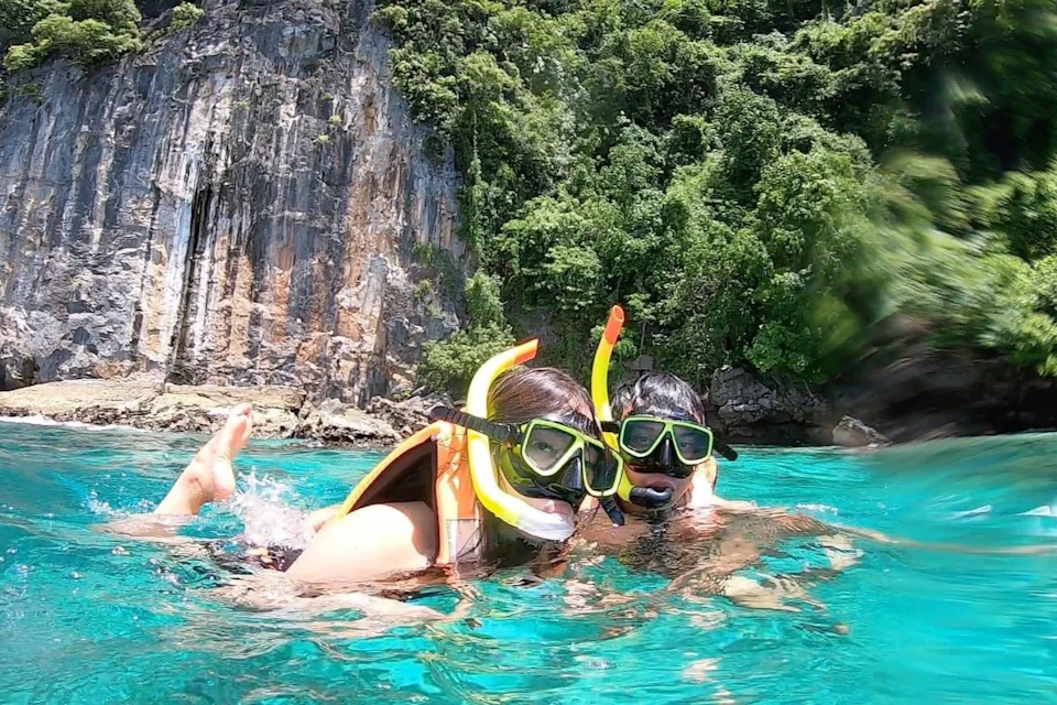 Two people snorkeling in clear turquoise water near a rocky, forested shoreline.