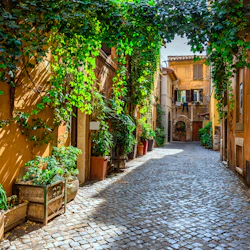 Smal kullerstensgata kantad av växter och murgröntäckta murar, vilket leder till rustika gula byggnader med balkonger och välvda dörröppningar.