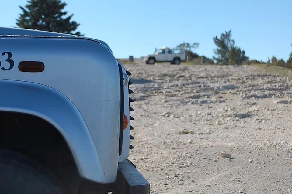 Front view of a silver vehicle with a rugged terrain and a white vehicle in the background under a clear blue sky.