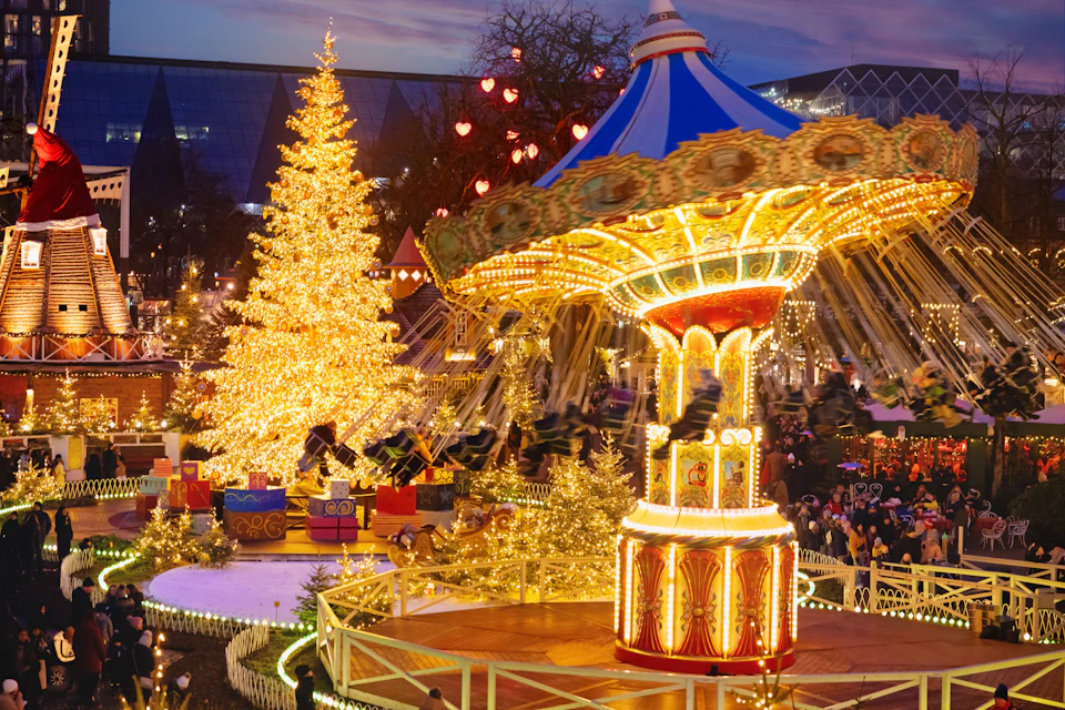 A vibrant carousel with colorful lights spins near an illuminated Christmas tree surrounded by festive decorations at night.