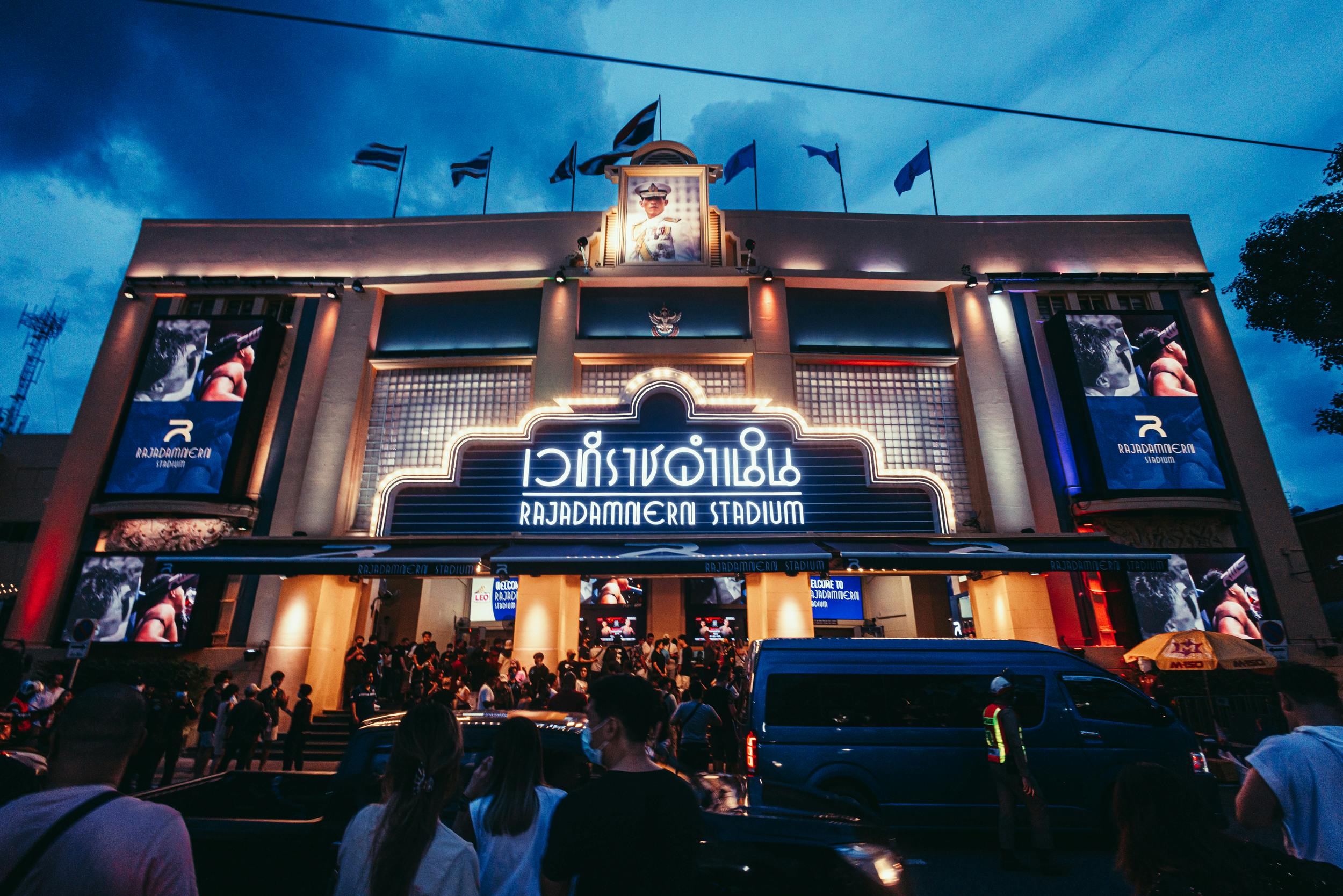 黄昏时分，一群人聚集在明亮的Rajadamnern体育场入口前，旁边有一张大海报和一辆蓝色面包车。