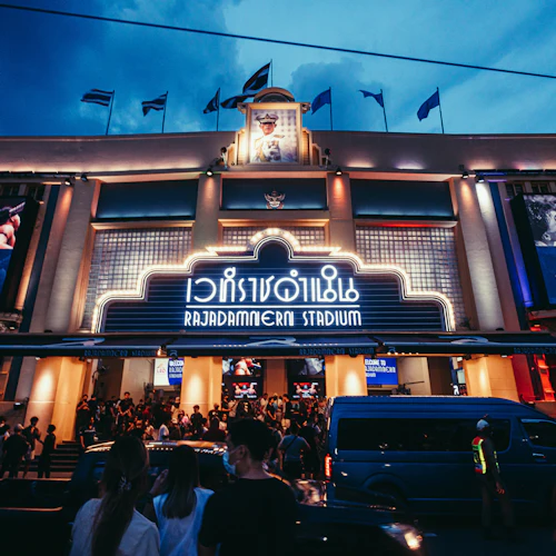 In der Abenddämmerung versammelt sich eine Menschenmenge vor dem hell erleuchteten Eingang des Rajadamnern-Stadions, in dessen Nähe große Plakate und ein blauer Lieferwagen stehen.