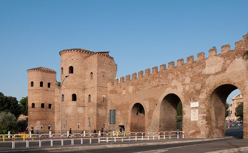 Ancient brick fortification with rounded towers and archways, under a clear blue sky; pedestrians and barriers in the foreground.