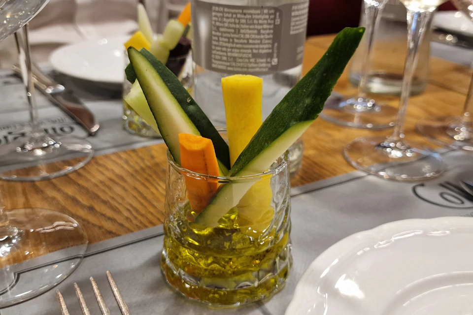 Vegetable sticks in a glass of olive oil on a table set for dining. Plates, forks, and glassware are visible in the background.