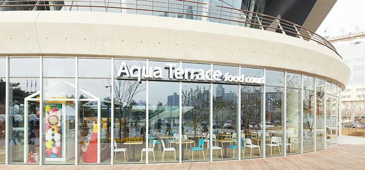 Widok z zewnątrz na strefę gastronomiczną Aqua Terrace, ze szklanymi ścianami, siedziskami na zewnątrz i pejzażem miejskim w tle.