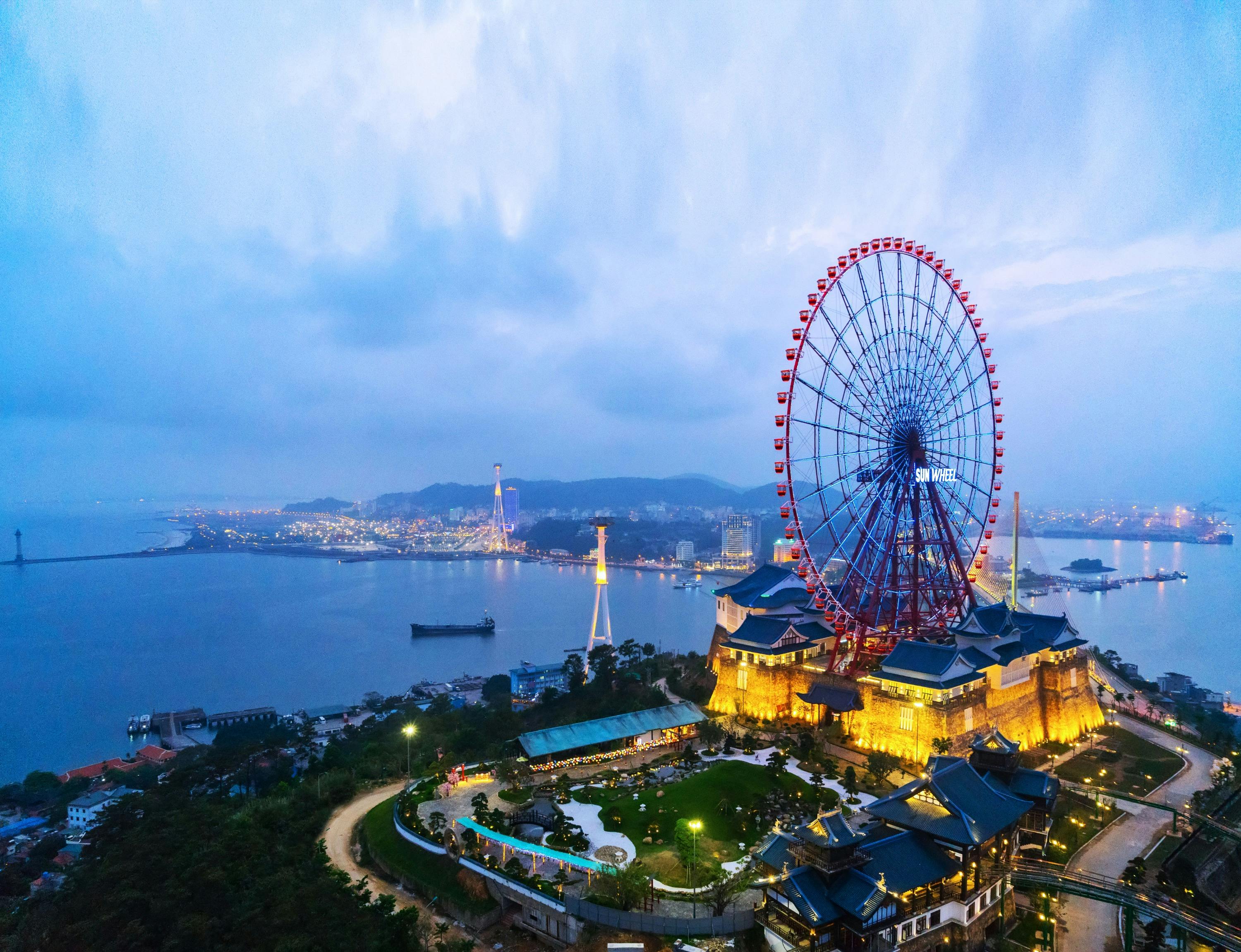 A brightly lit ferris wheel near a body of water, surrounded by illuminated buildings and cityscape at dusk.