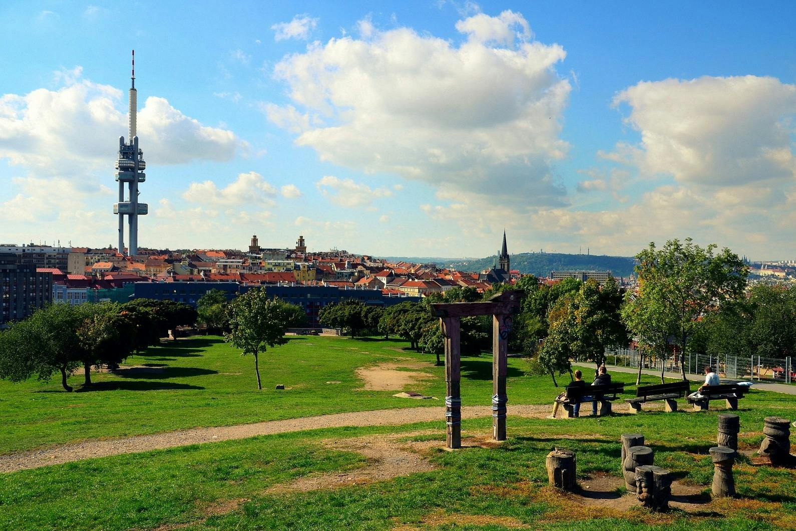 Un parc avec de l'herbe verte, des arbres, un sentier et des bancs. Un paysage urbain avec des clochers d'église est visible sous un ciel partiellement nuageux.