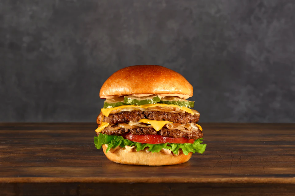 A cheeseburger with two beef patties, lettuce, tomato, onions, pickles, and cheese, on a wooden surface against a dark background.