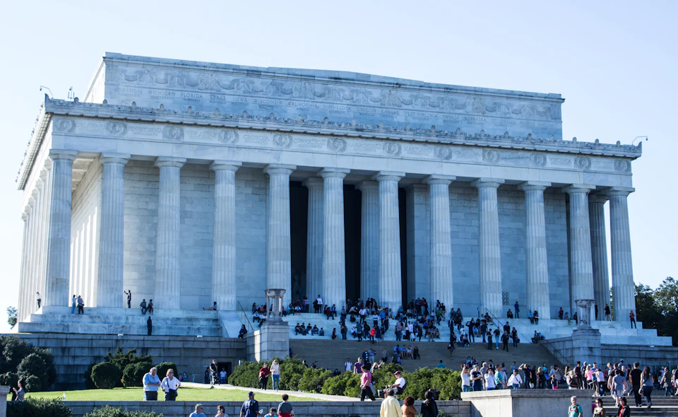 The image shows a large neoclassical building with columns, with many people gathered on its steps and lawn under a clear sky.