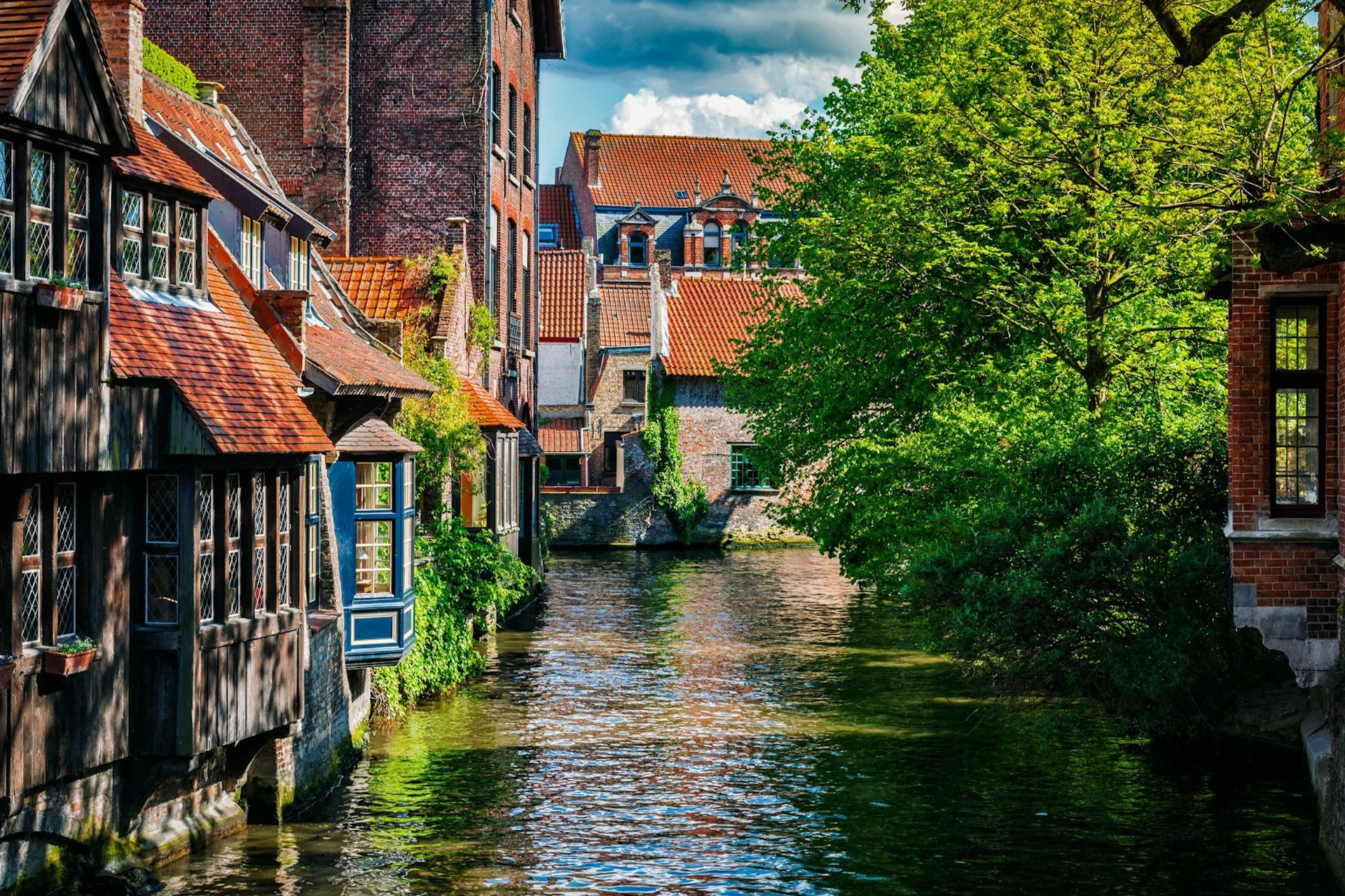 A canal flanked by brick buildings with red-tiled roofs on the left and lush trees on the right, under a partly cloudy sky.
