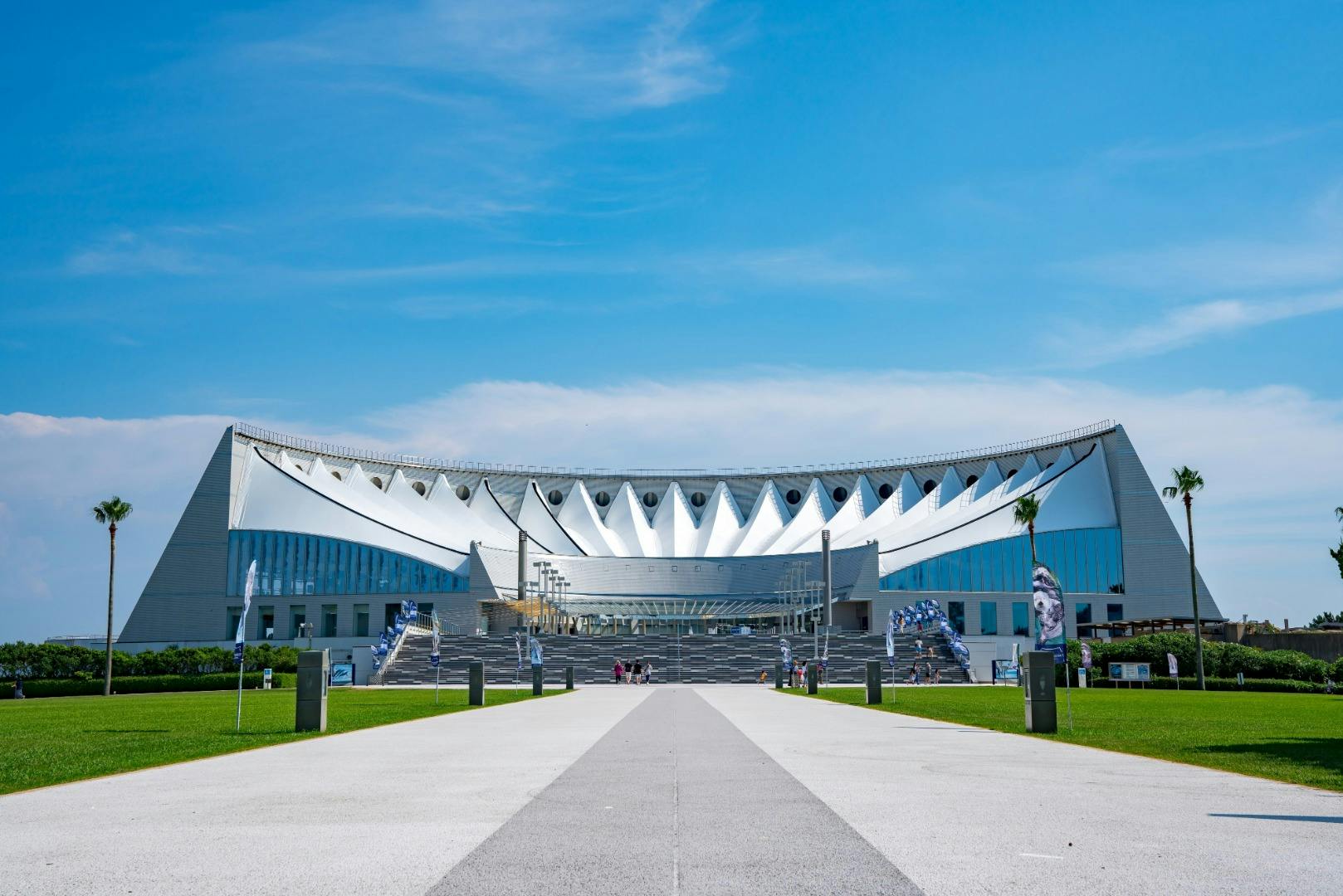Un stade moderne au toit blanc et à la forme triangulaire, vu depuis une allée bordée de gazon et de drapeaux, sous un ciel bleu.