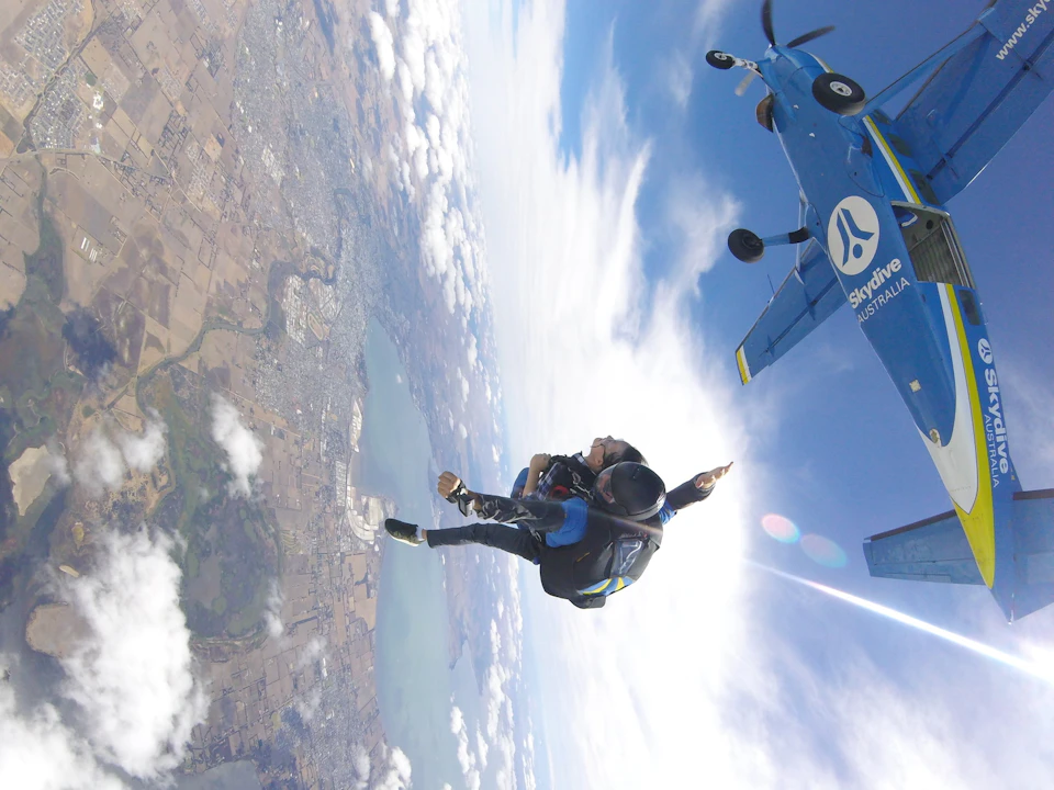 Two people tandem skydiving with a "Skydive Australia" plane in the background, high above a landscape with fields and a body of water.