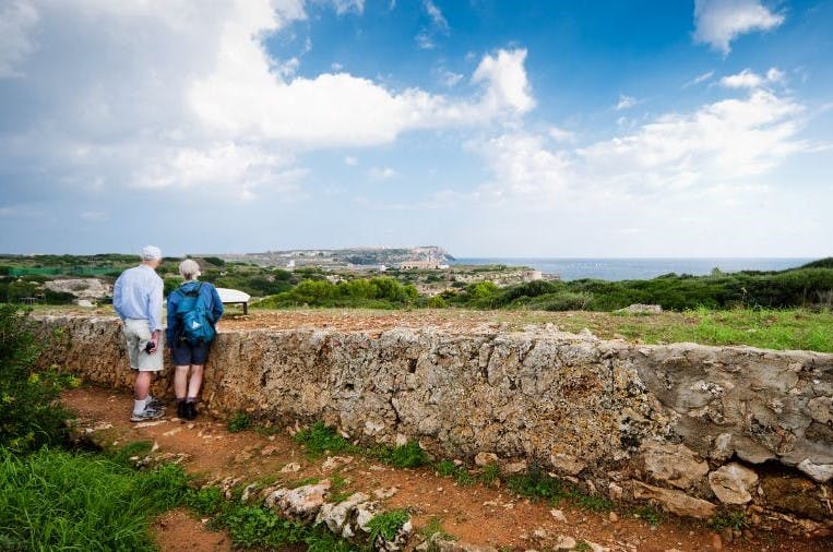 Δύο άνθρωποι στέκονται πίσω από έναν πέτρινο τοίχο, με θέα ένα γραφικό τοπίο με πράσινο και μια μακρινή ακτογραμμή κάτω από έναν εν μέρει συννεφιασμένο ουρανό.