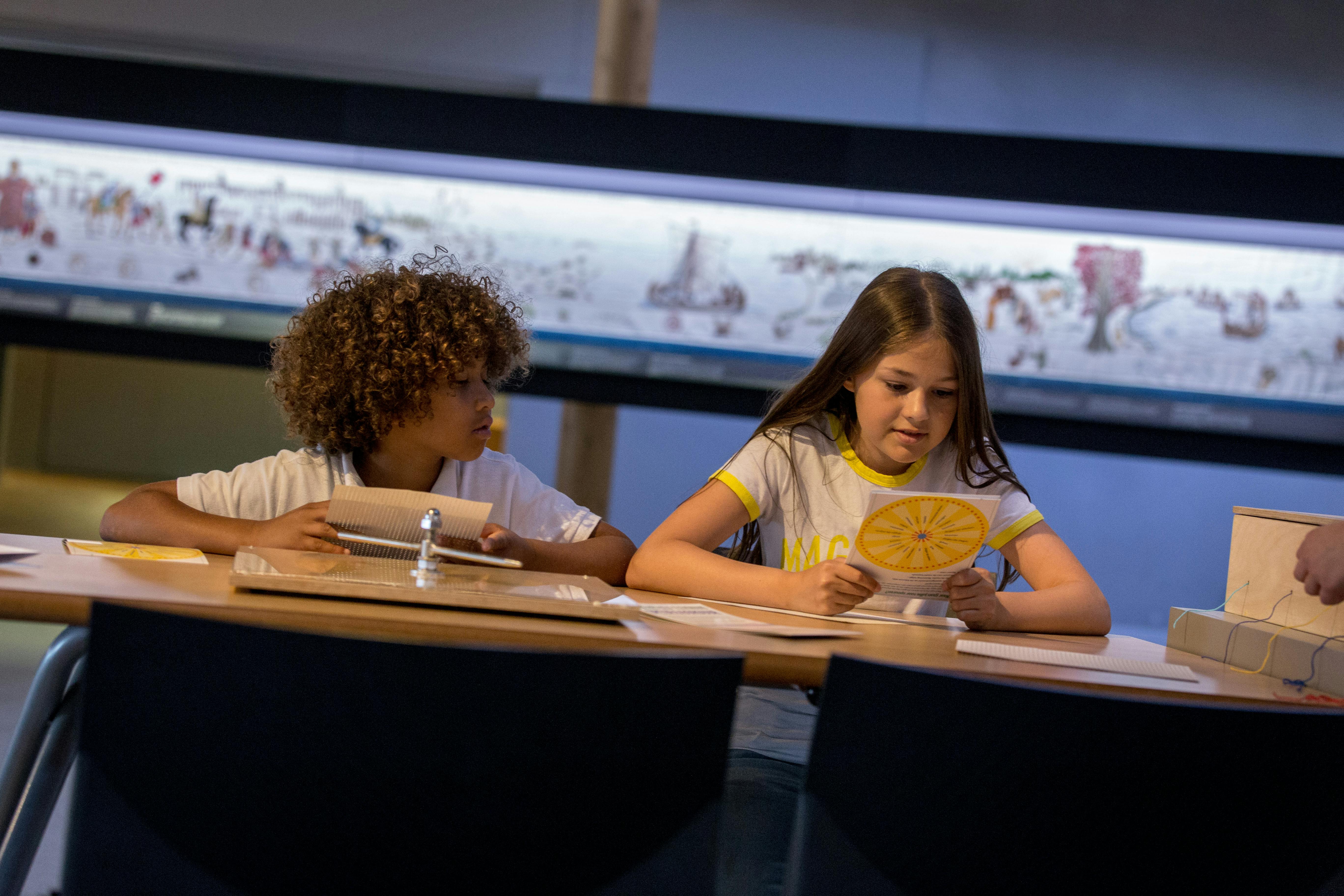 Děti sedí u stolu a zkoumají vzdělávací materiály. V pozadí je vidět velký displej s kresbami nebo kresbami.