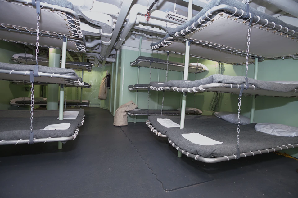 A room with stacked metal bunk beds and gray mattresses; some bags hang on the walls.