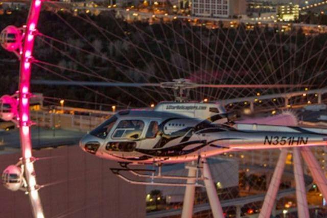 A helicopter in flight near an illuminated cityscape, with a large observation wheel visible in the background.