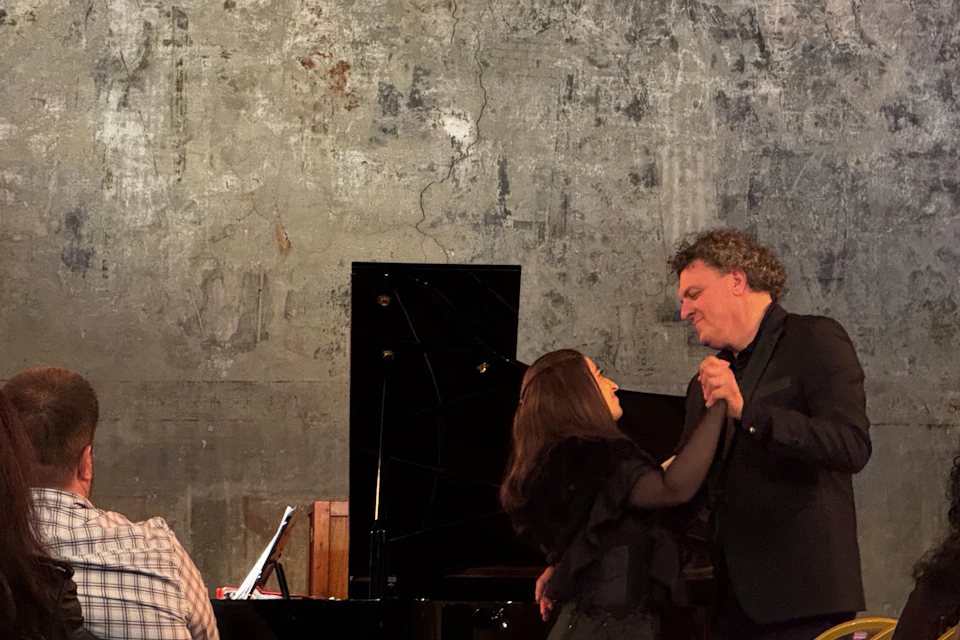 A man and a woman are dancing in front of a grand piano, with a textured wall in the background.