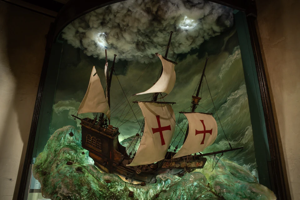 Wooden ship with large sails, featuring red crosses, navigating rough green waters under a cloudy sky.