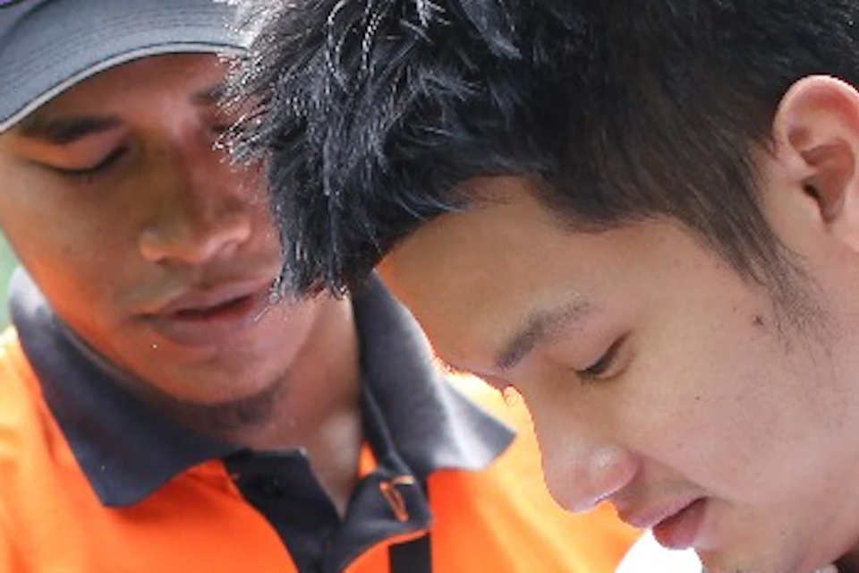 Two individuals are preparing for an outdoor activity, one assisting the other with safety equipment near a fence in a lush park.