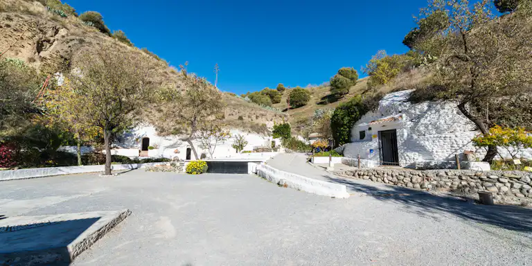 Habitations troglodytes aux murs blancs et arbres dans un paysage sec et vallonné sous un ciel bleu clair. Ombres de personnes au premier plan.