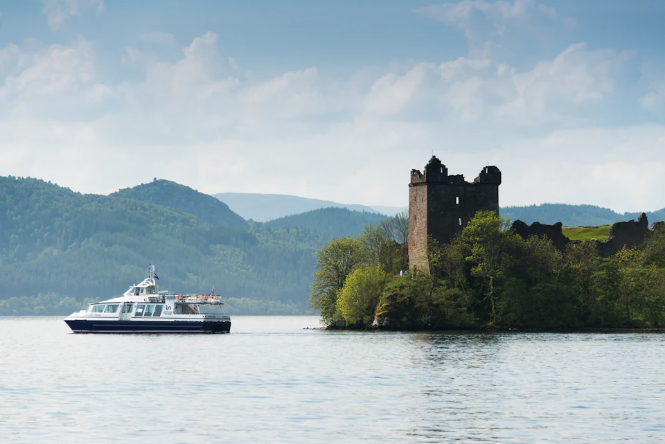 A boat sails near a historic, partially ruined castle on a small, tree-covered island amidst a tranquil lake, with forested hills in the background.