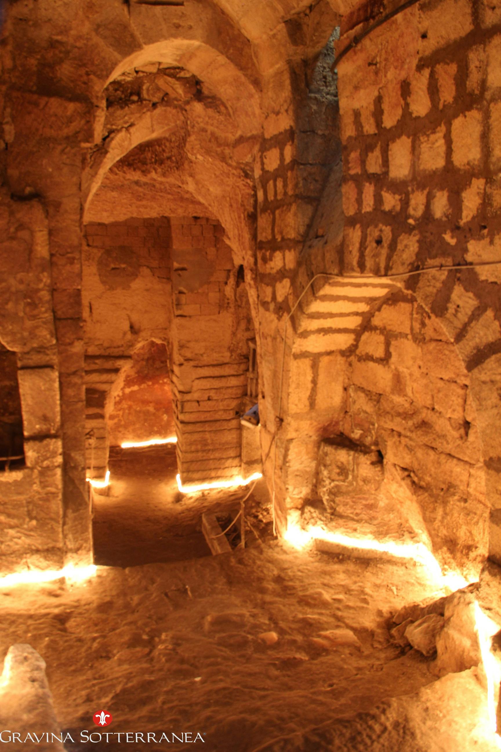 A well-lit, ancient stone underground passage with arched doorways and walls. Dim, warm lighting emphasizes the architectural details.