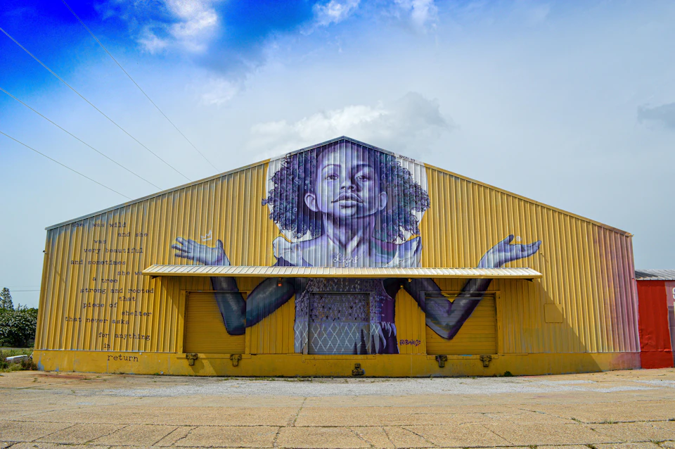 A large mural of a child with curly hair and outstretched arms is painted on a yellow warehouse under a partly cloudy sky.