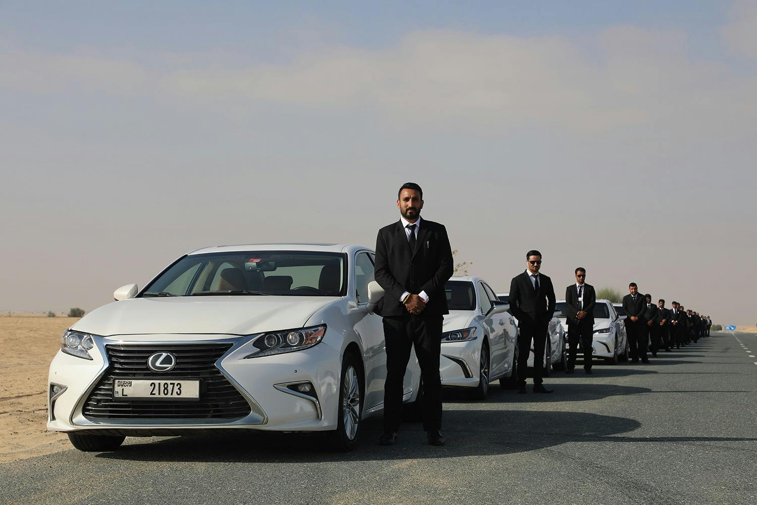 Män i svarta kostymer står bredvid en rad vita bilar på en öppen väg, som sträcker sig i fjärran under en delvis molnig himmel.