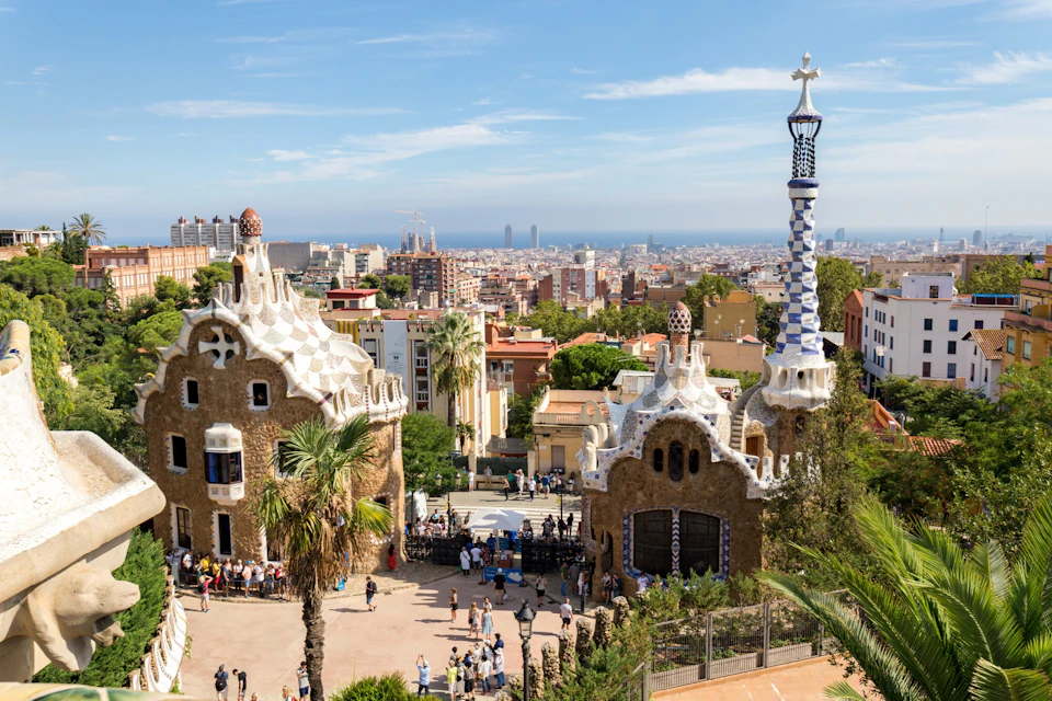 A view of colorful buildings and a tower in Park Güell, Barcelona, with the cityscape and sea in the background.
