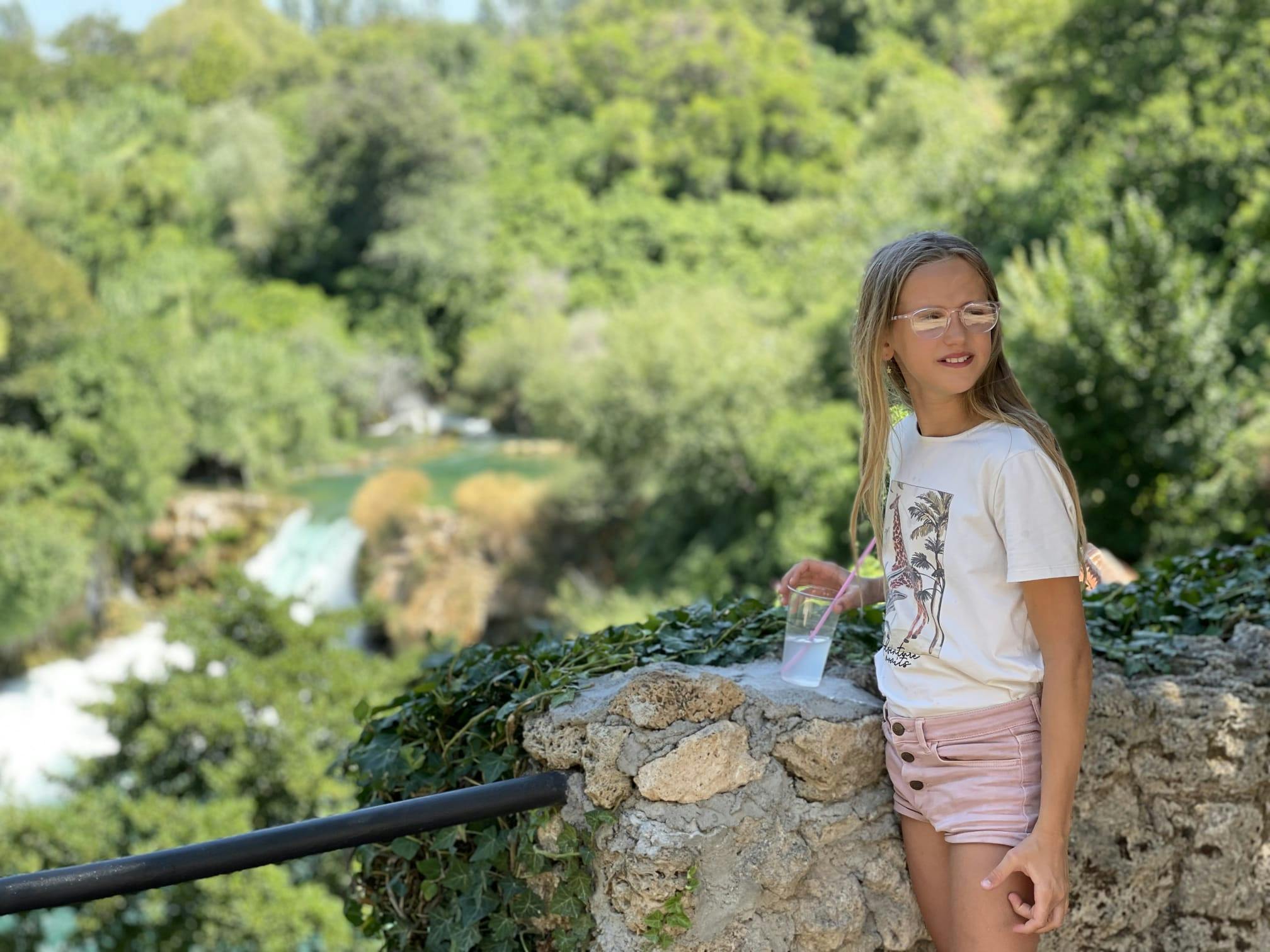 一位戴着眼镜的女孩拿着饮料，站在石墙旁，背景是郁郁葱葱的绿色植物和瀑布。