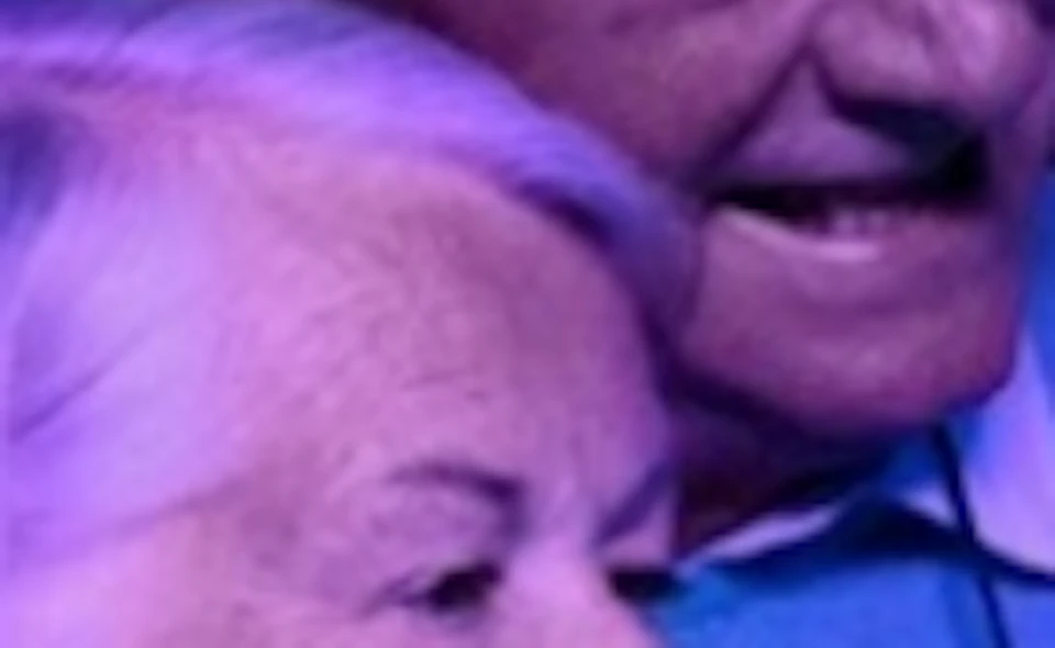 An elderly couple smiles while looking at a jellyfish exhibit under purple lighting.