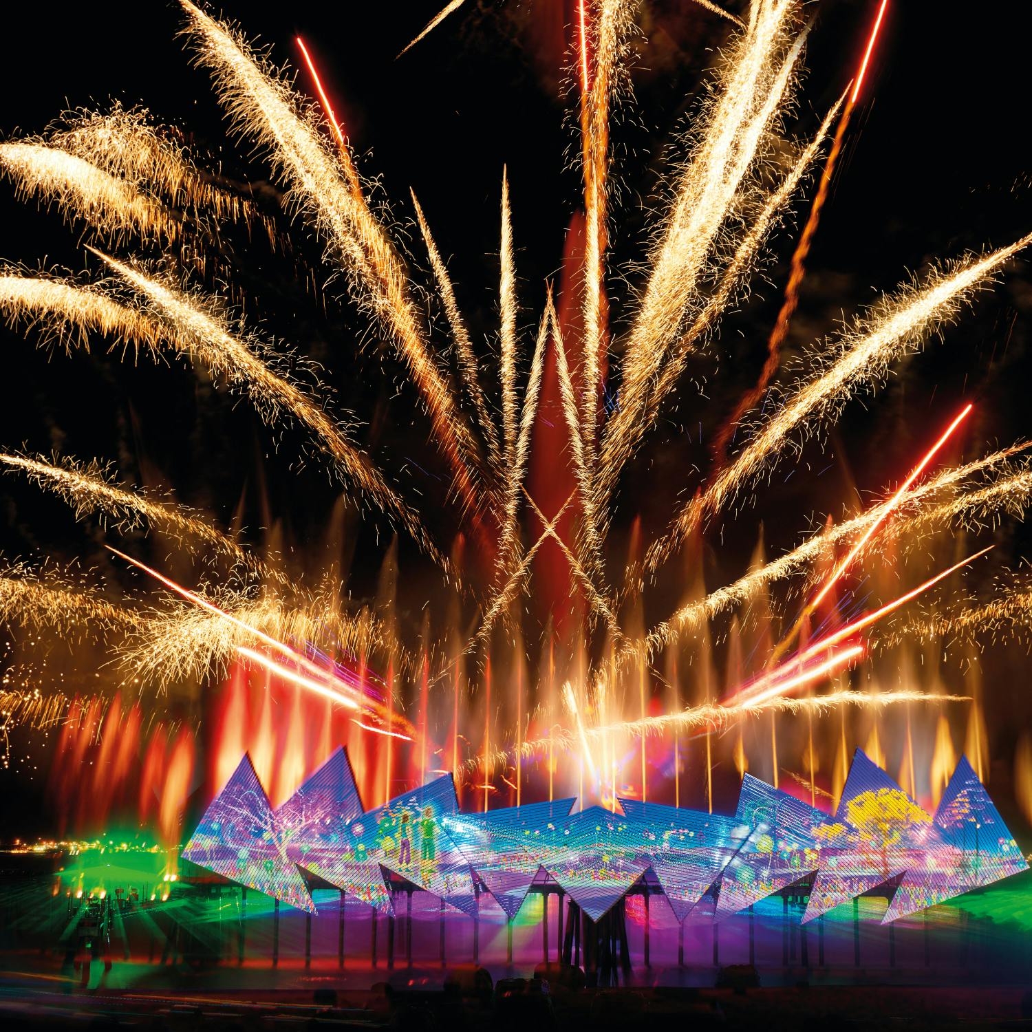 Barevný ohňostroj exploduje na noční obloze nad osvětleným geometrickým jevištěm se zářivými světly a vzory.