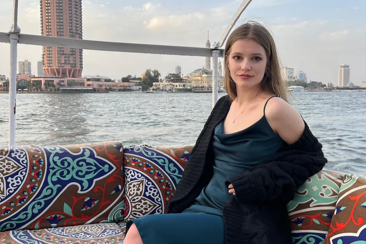 一位身穿蓝色连衣裙和黑色毛衣的女士坐在船上，船上有五颜六色的靠垫，背景是城市景观和水体。