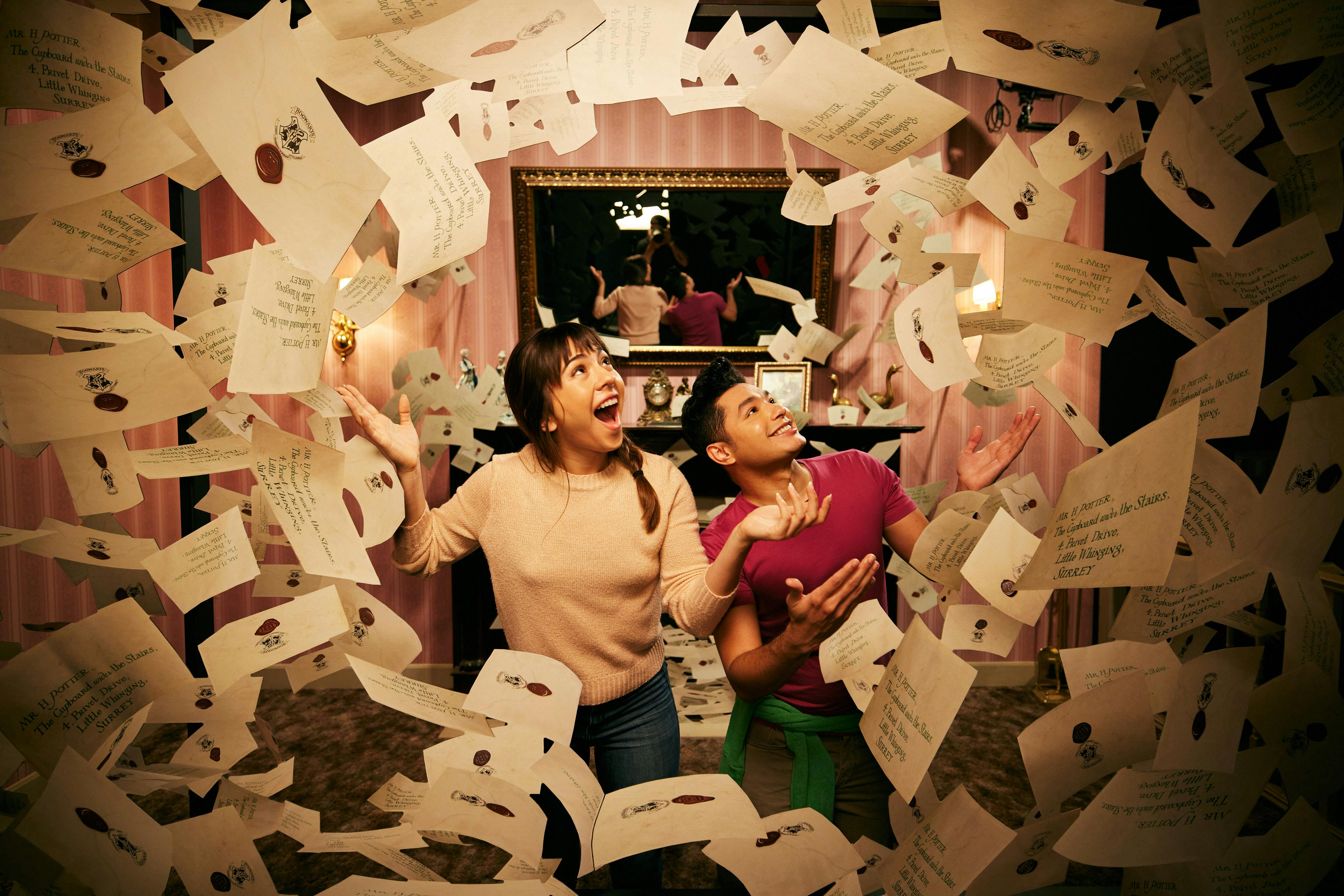 Two people joyfully surrounded by flying envelopes in a decorated room with a mirror and pink wallpaper.
