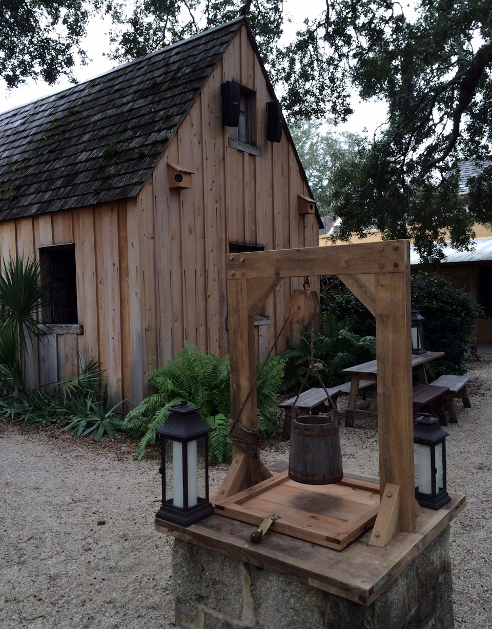A wooden well structure with lanterns stands in front of a rustic wooden cabin surrounded by plants and trees.