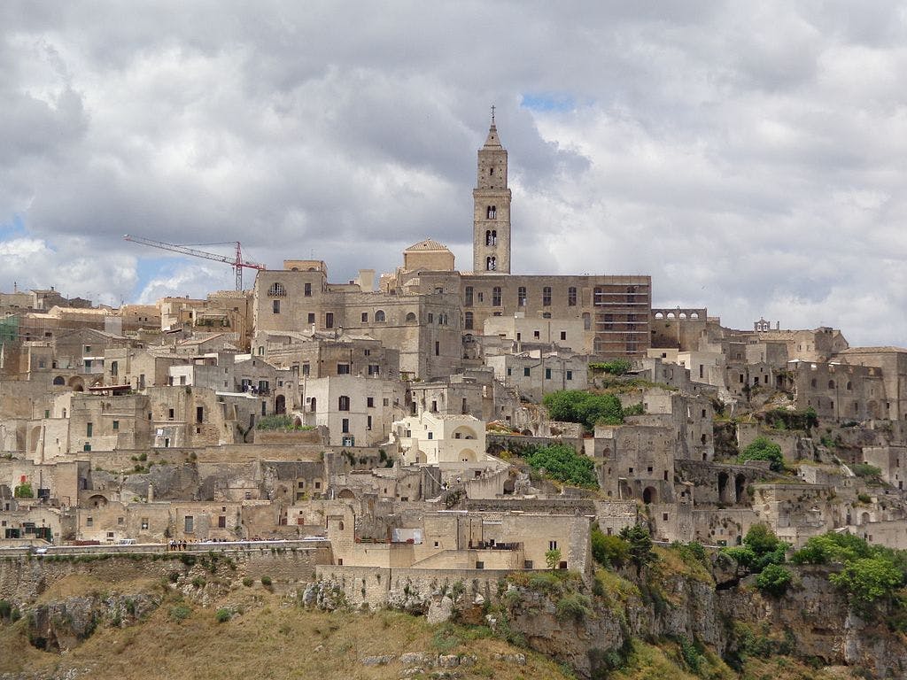 A hillside town with closely packed stone buildings, a tall church tower, lush greenery, and clouds overhead.